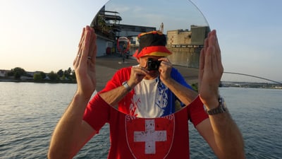 Mann hält runden Spiegel vor Gesicht am Wasser.