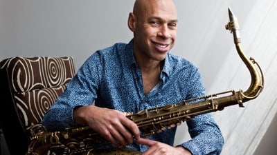 Künstler mit Saxophon auf braunen Sessel sitzend, lächelnd. 