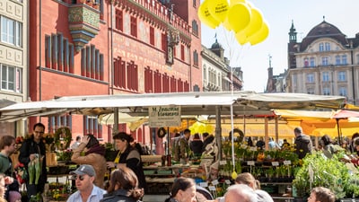 Menschen sitzen an Tischen auf einem Platz vor einem roten Gebäude mit Ballons und Marktständen.