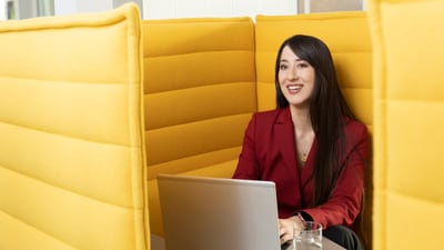 Frau arbeitet lächelnd an einem Laptop in einer gelben, gepolsterten Kabine.