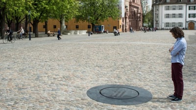 Person steht auf dem Platz vor dem Basler Münster und betrachtet die kreisrunde Gusseisenplatte im Plastersteinboden, welche den römischen Sodbrunnen markiert