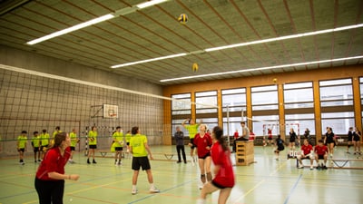 Volleyballspiel in einer Turnhalle mit Spielern in roten und grünen Trikots.