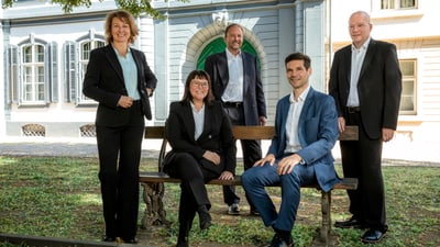 Fünf Personen in formeller Kleidung vor historischer Architektur sitzend und stehend.