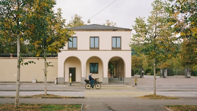 Person auf Fahrrad vor zweistöckigem Gebäude und Bäumen.