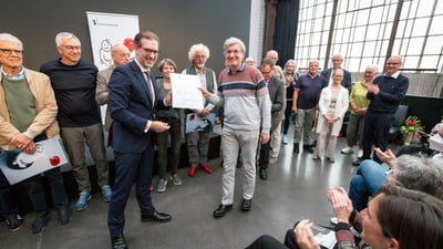 Mann erhält Auszeichnung, von einer Gruppe umgeben, während Publikum applaudiert.