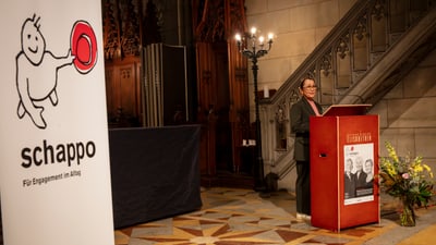 Rednerin am Podium neben Schappo-Banner in festlicher Halle.