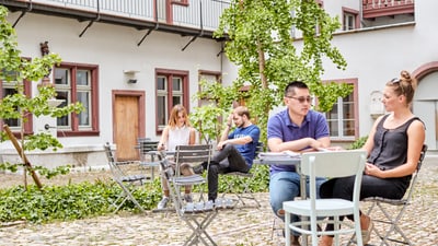 Menschen sitzen an Tischen im gepflasterten Innenhof mit Pflanzen und alten Gebäuden.