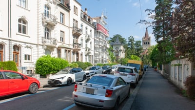 Strasse mit geparkten Autos und Gebäuden im Hintergrund, Kirchturm sichtbar.",