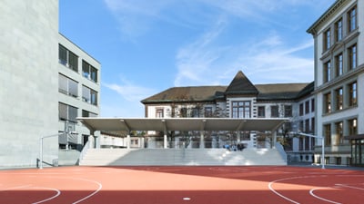 Schulhof mit rotem Sportplatz und einer Tribüne hin zum Platz.