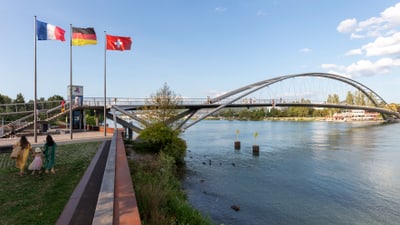 Foto der Dreiländerbrücke im Sommer, links wehen die Fahnen von Frankreich, Deutschland und der Schweiz im Wind.