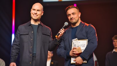 Zwei Männer auf einer Bühne mit Mikrofon und Auszeichnung.