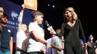 Joelle Perrret interviewt einen Schüler auf Bühne, umgeben von weiteren Schülerinnen.