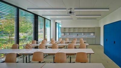 Moderner Klassenraum mit Holztischen und Stühlen, Aussicht auf Bäume durch Fensterläden.