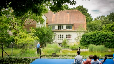 Personen entspannt auf blauer Matte im Garten vor einem Haus.