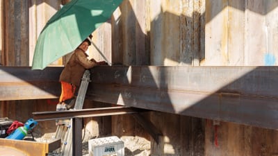 Arbeiterin auf Leiter unter grünem Sonnenschirm bei Baustelle.",