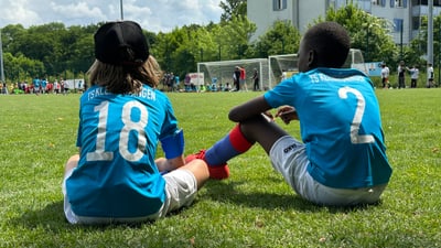 Zwei Kinder in Fussballtrikots sitzen auf dem Rasenplatz am Spielfeldrand.