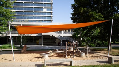 Kinderspielplatz mit orangefarbenem Sonnensegel und Gebäude im Hintergrund.