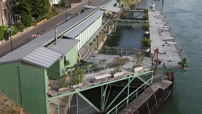 Grüne Garderobengebäude am Rheinufer, vor denen sich ein Steg auf dem Rhein befindet.