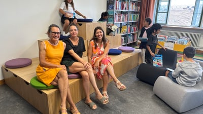 Caroline Stauffer, Michèle Baitsch und Sara Gysin in einer Bibliothek sitzen auf Holzstufen mit Bücherregalen im Hintergrund.
