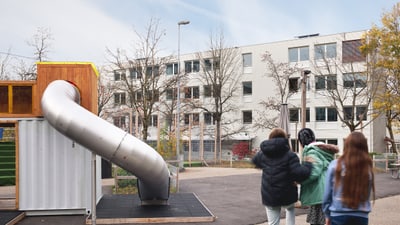 Mehrere Kinder auf einem Spielplatz mit Rutsche und Gebäude im Hintergrund.