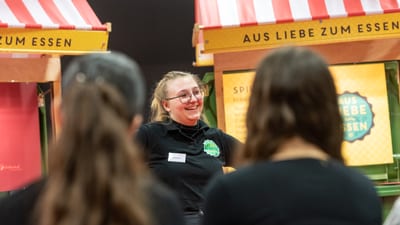 Lächelnde Frau spricht zu Publikum, Marktstände im Hintergrund.