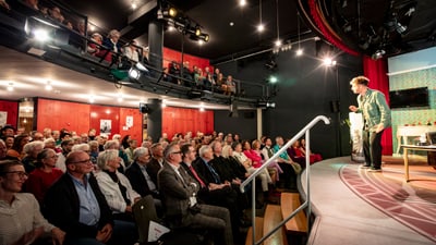 Person spricht vor Publikum in Theater mit Galerie.",