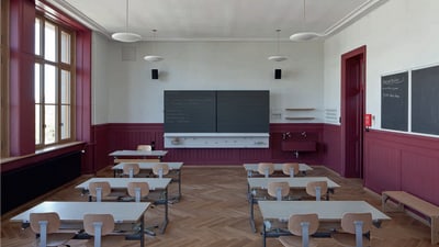 Klassenzimmer mit hölzernen Tischen, roten Wänden und Tafel.