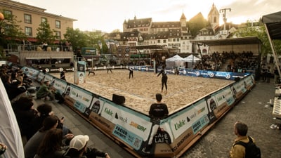 Beachvolleyball-Spiel im Stadtzentrum mit Publikum und historischen Gebäuden im Hintergrund.
