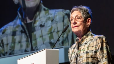 Frau hält Rede auf einer Bühne mit Mikrofon und Podium, Hintergrundbild derselben Frau.