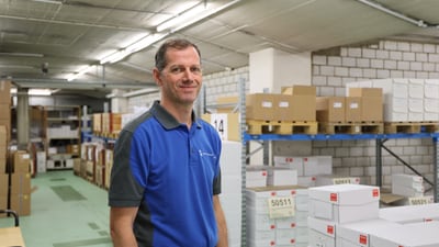 Mann in blauem Hemd steht in einem Lagerraum mit gestapelten Kartons.