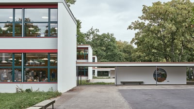 Zweistöckiges Schulhaus, davor ein Platz und ein Unterstand mit einer Sitzbank.