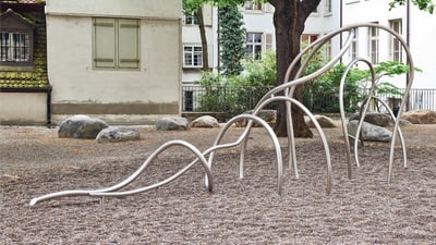 Kiesplatz mit verschlungenen Metallstangen und grossen Steinen mit einem Baum.