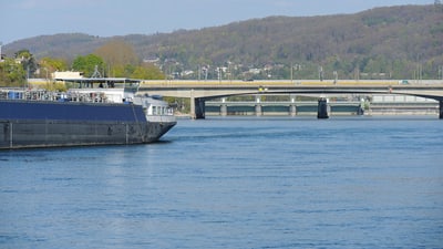 Schiff auf dem Rhein mit Brücke und bewaldeten Hügeln im Hintergrund.