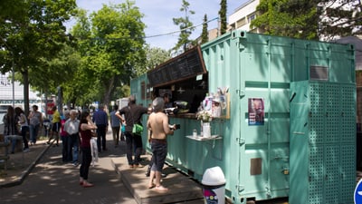 Essensstand in einem umgebauten Container mit Warteschlange von Menschen im Freien.