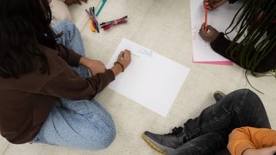 Personen sitzen auf dem Boden und zeichnen auf Papier mit Buntstiften.