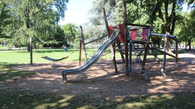 Zwischen grossen Bäumen steht ein Spielplatz mit Kletterhaus und Rutschbahn