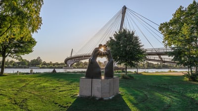 Skulptur vor einer Fussgängerbrücke bei Sonnenuntergang über einem Fluss.