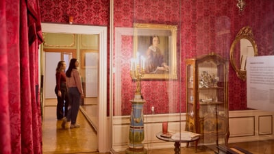 Zwei Personen betrachten ein historisches Zimmer mit roten Wänden und Gemälden.
