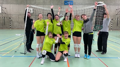 Jubelnde junge Frauen in Sporttrikots, halten einen Pokal in einer Turnhalle hoch.