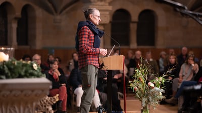 Person hält eine Rede vor Publikum in einem Saal.