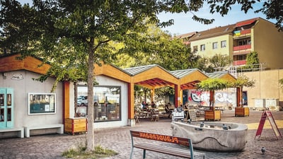 Öffentlicher Platz mit Bänken, Bäumen und Cafés bei Sonnenschein.