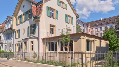 Dreistöckiges Haus mit grünem Fensterläden und Gartenzaun bei blauem Himmel.
