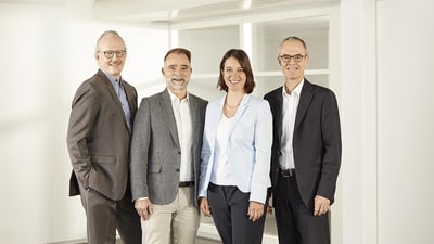 Portrait der AWA-Geschäftsleitung. Von links nach rechts sind Dieter P. Wirth, Michael Mauerhofer, Nicole Hostettler und Samuel Hess nebeneinanderstehend zu sehen. Alle lächeln freundlich und offen in die Kamera und tragen Anzüge.