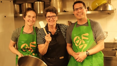 Drei Personen in einer Küche mit Schürzen und Kochutensilien.