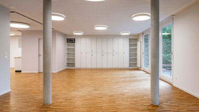 Leerer Raum mit Holzboden und weissen Wänden, zwei Säulen, Einbauschränken und grossen Fenstern.