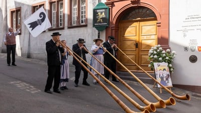 Menschen in traditioneller Kleidung spielen Alphorn vor einem Gebäude.