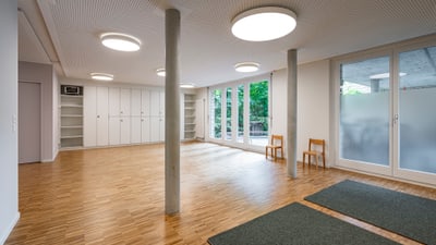 Heller Raum mit Holzboden, Säulen und Fenstern, zwei Stühle an der Wand.