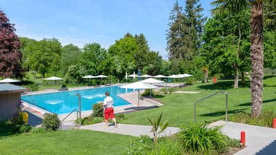 Freibad mit grünem Rasen und Sonnenschirmen, Person geht vorbei.