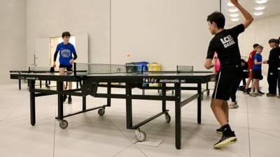 Junge spielt Tischtennis in einer Halle mit Zuschauern im Hintergrund.