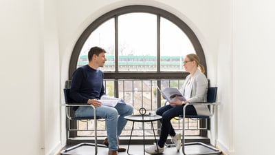 Zwei Menschen sitzen in modernen Stühlen vor einem grossen Fenster und unterhalten sich.